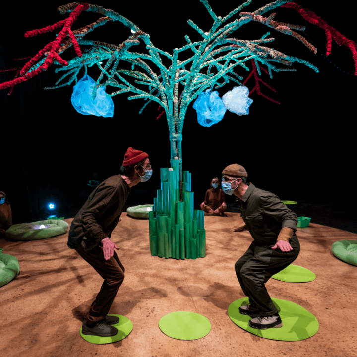 actors face each other with bent knees. They are standing on green circles on the floor. The green tree is in the background of the stage.