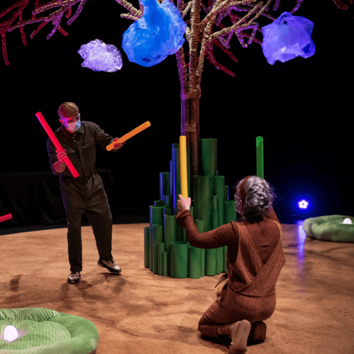 Two actors holding colour sticks. One is kneeling with their back to the camera in front of the tree.