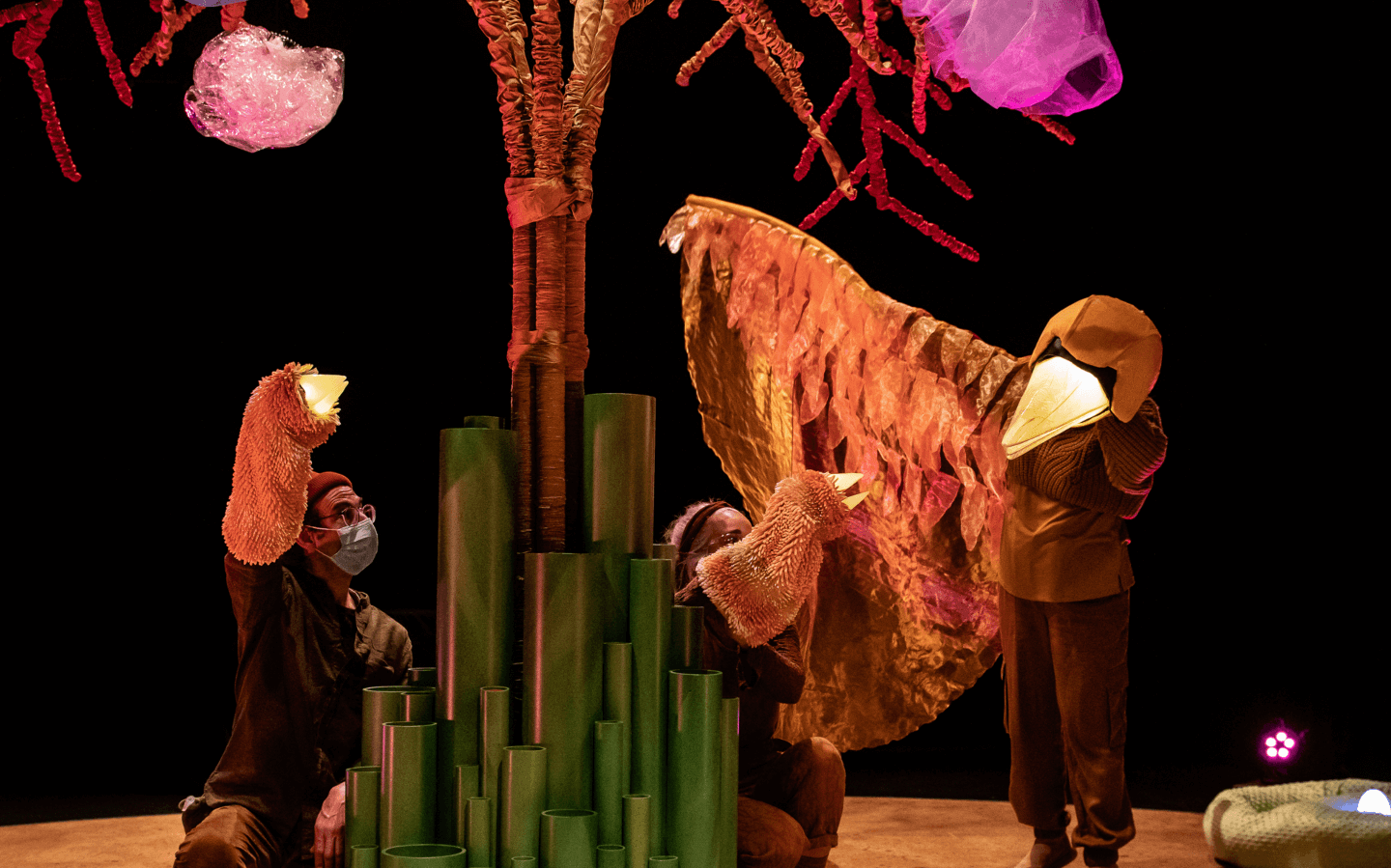 3 actors gather on stage with puppets under an artistic tree softly lit. The puppets are brown birds with their beaks lit up. One of the birds is larger and is extending a wing towards the two smaller birds.