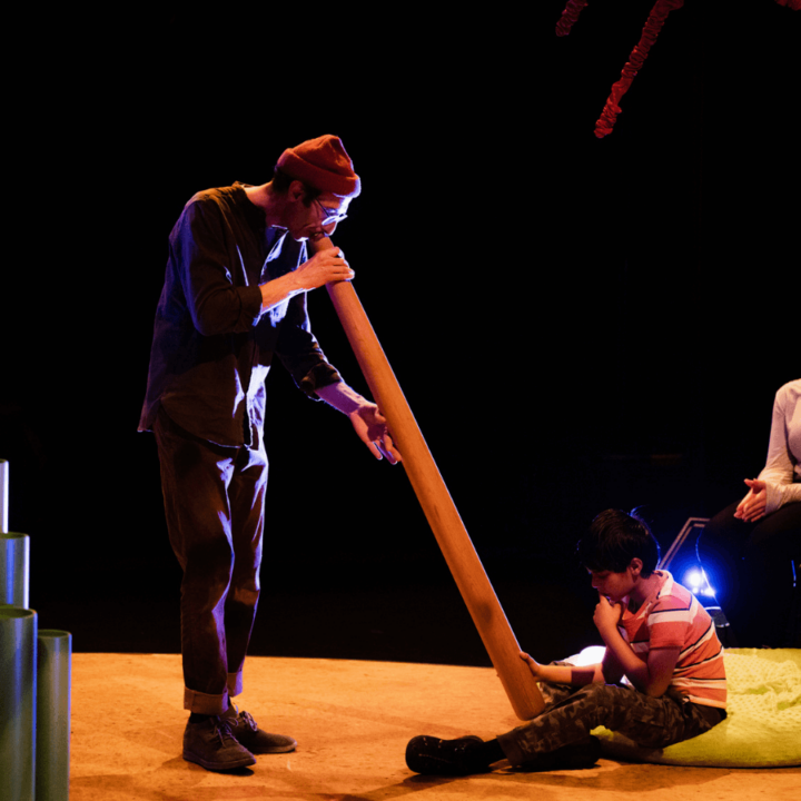 A performer plays a didgeridoo while a child sitting on stage curiously inspects the instrument.