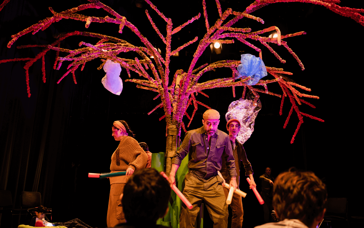 Four performers are gathered around a tree in the centre of the stage, holding drum sticks.