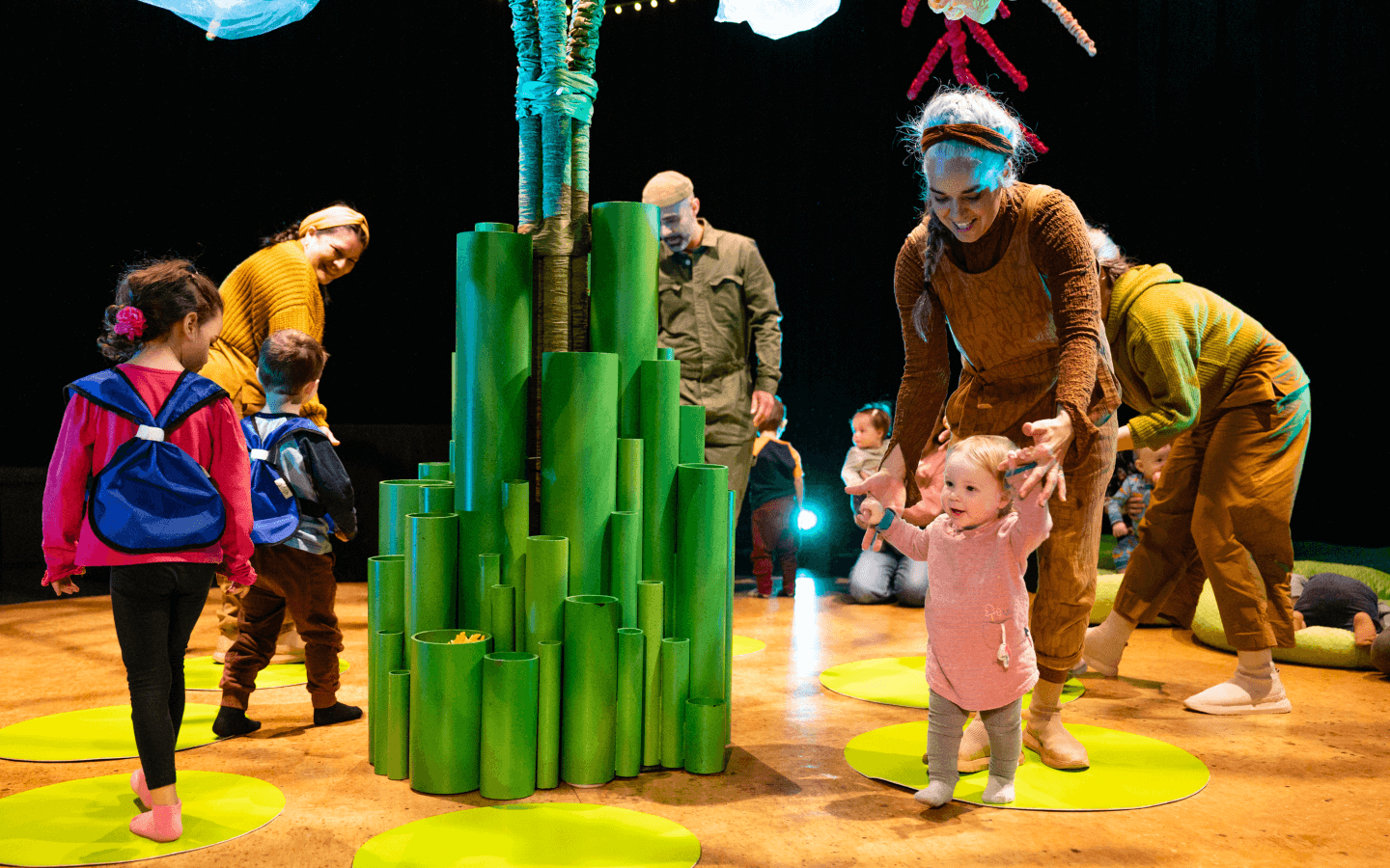 A performer helps a small child in the on-stage activity. The performer, a few children, and a few parents, all walk around a tree in the centre of the stage.