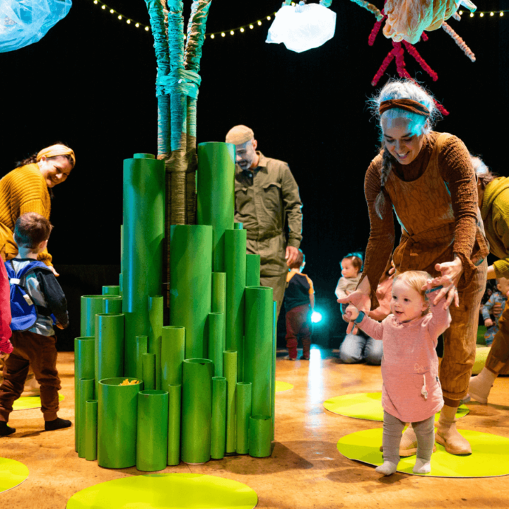 A performer helps a small child in the on-stage activity. The performer, a few children, and a few parents, all walk around a tree in the centre of the stage.