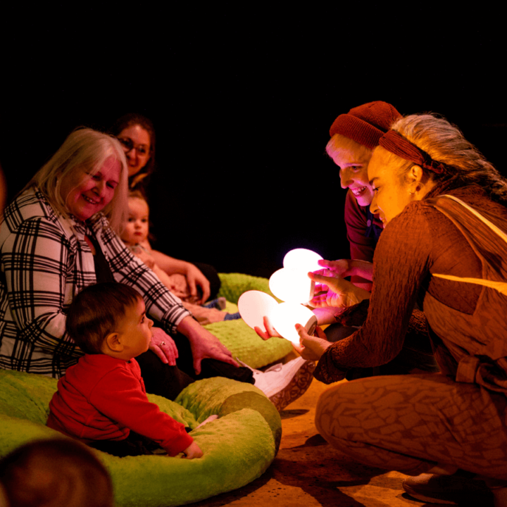 Two small children sitting in bean bags with their guardians, and two performers from Tree. They're all gathered around and observing small light-up eggs.