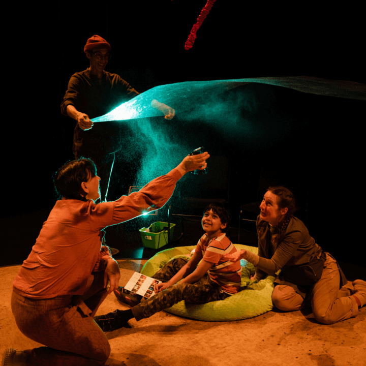 A child and their parent watches in awe as two performers create magical visuals using a scarf, and coloured lights above their heads.