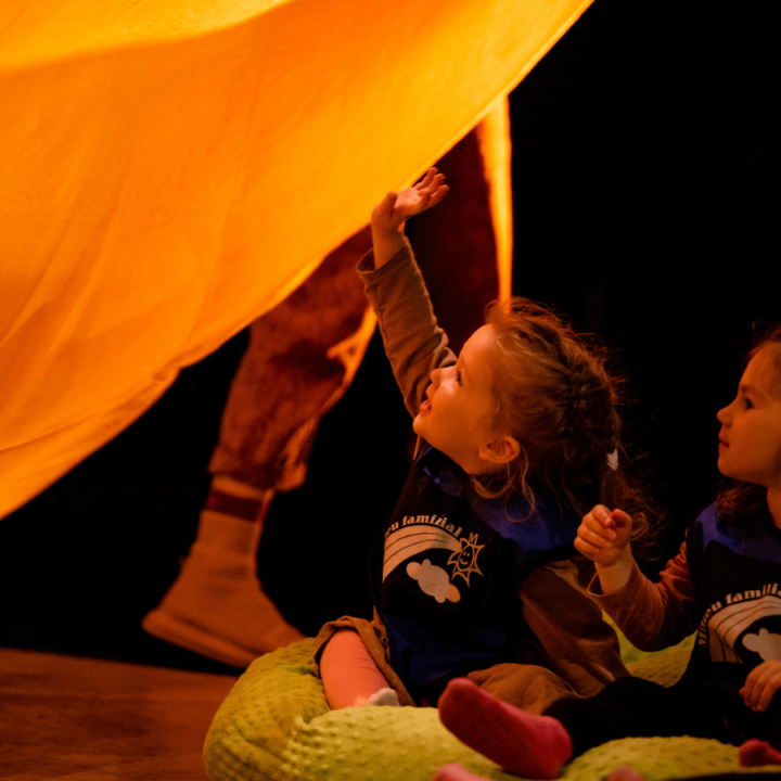 A small girl sitting on stage with a friend in a bean bag, smiling, and reaching up to touch a large yellow tapestry.