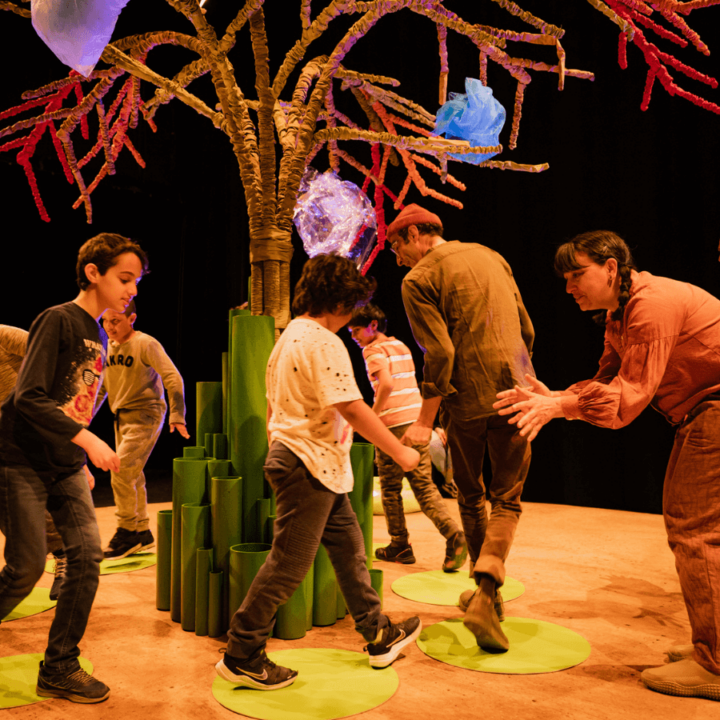 Two performers, some parents, and their children all dance in a circle around a tree on stage.