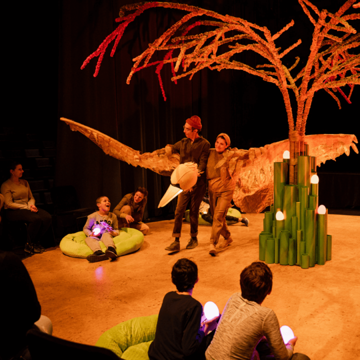 Two performers walk around centre stage, surrounded by audience members who sit in a circle around them. The performers link arms and hold out two large wings to either side; to look like a single, large, bird puppet flying around a tree.