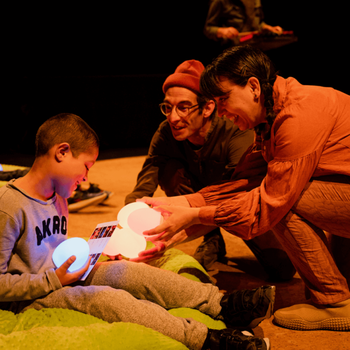 Two performers observe a handful of colourful and luminous toy eggs - letting a child in the audience play with them.