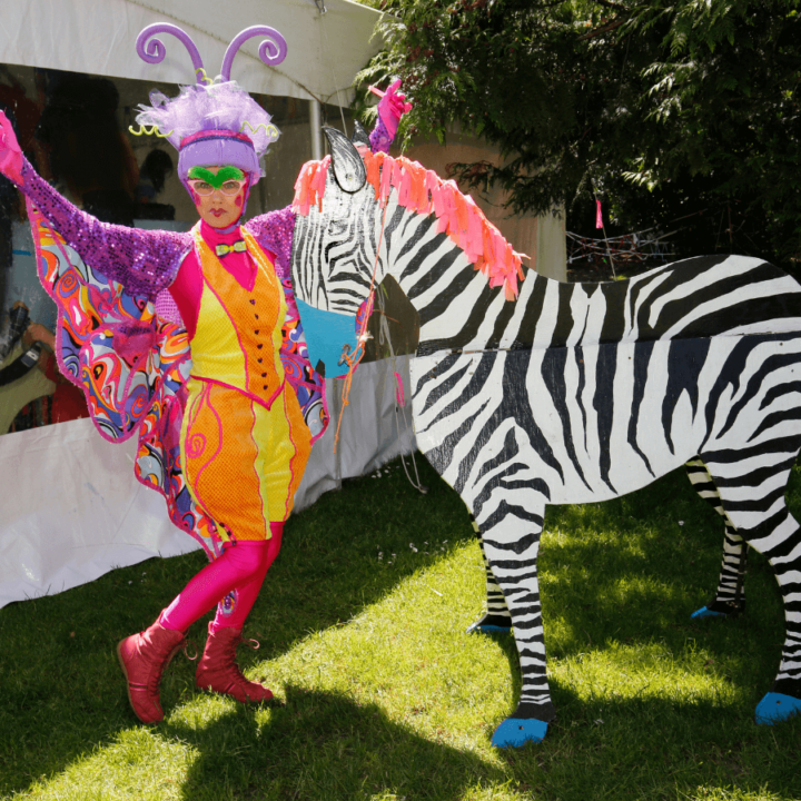 A picture of a roaming performer, dressed in colourful make-up and a butterfly costume, posing with a wooden cutout of a zebra.