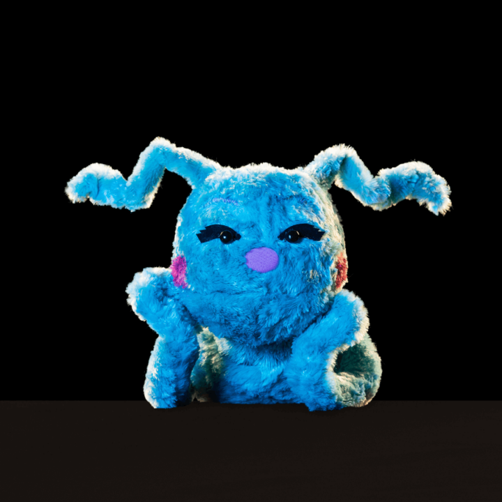 A promotional image for the show "Division Infinity Saves the World" - displaying a blue puppet, with antennae hair, named Chiqui. The puppet poses with their head resting in their hands.
