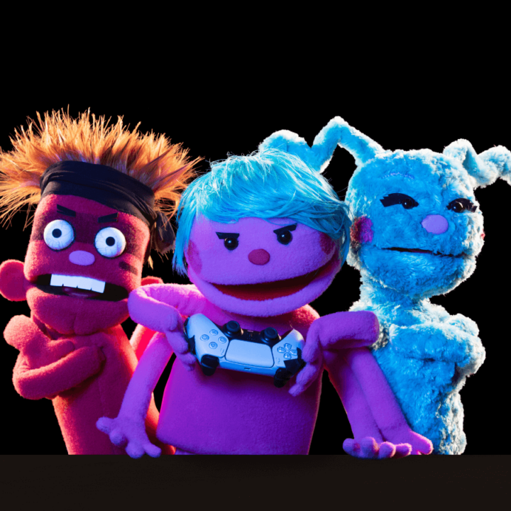 A promotional image for the show "Division Infinity Saves the World" - displaying their three new puppets, Spiderwolf, Chiqui, and Soda. Spiderwolf, the red puppet on the far left of the image has crazy spiked hair and is wearing a sweatband The middle puppet, Soda, poses with her arms around her fellow puppets. Chiqui, the blue puppet on the far right, hugs Soda.