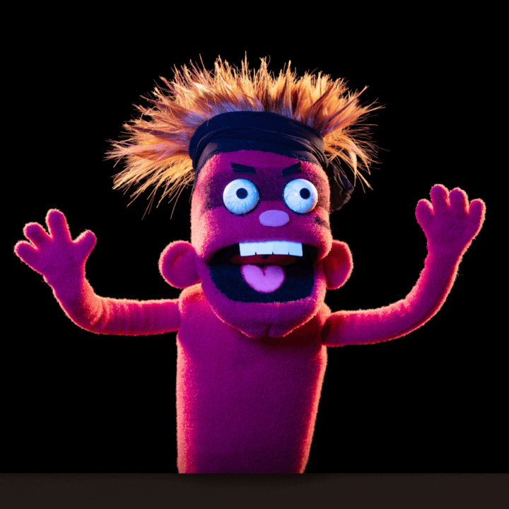 A promotional image for the performances of "Division Infinity Saves the World". This image shows their puppet, named Spiderworlf, posing with their arms up in the air, as if they were growling. This puppet is red, with spiked red hair, and is wearing a karate headband.