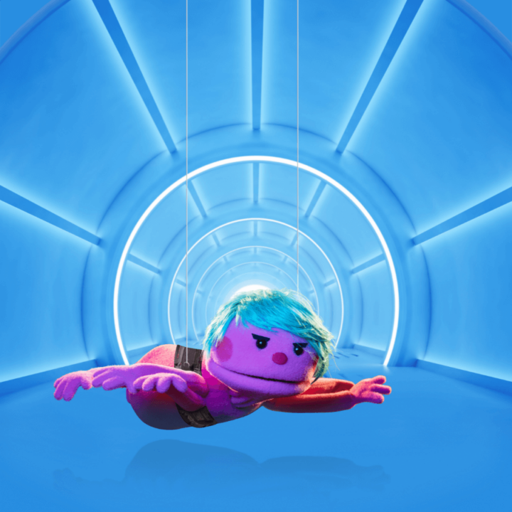 A promotional image for the show "Division Infinity Saves the World" - displaying a purple puppet, with blue hair, named Soda. Soda is posing "mission impossible style" - suspending and grappling down from the ceiling like a secret agent.