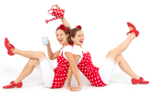 Twin women sitting back to back in red polka dot bathing suits and red shoes. One is pretending to pour tea over her head into the other woman's cup.