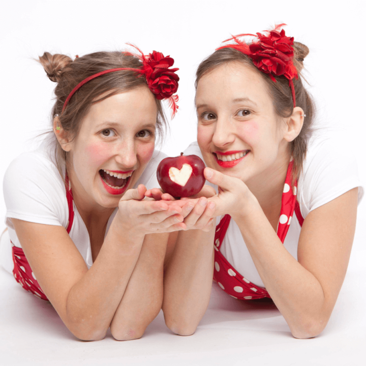 Twin women holding an apple together with a bite in it that looks like a heart. The women are wearing red and white.