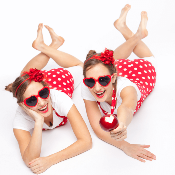 Twin women are lying beside each other looking up at the camera. One is holding an apple with a bite in it that looks like a heart. The women are wearing red and white and red heart shaped sunglasses.