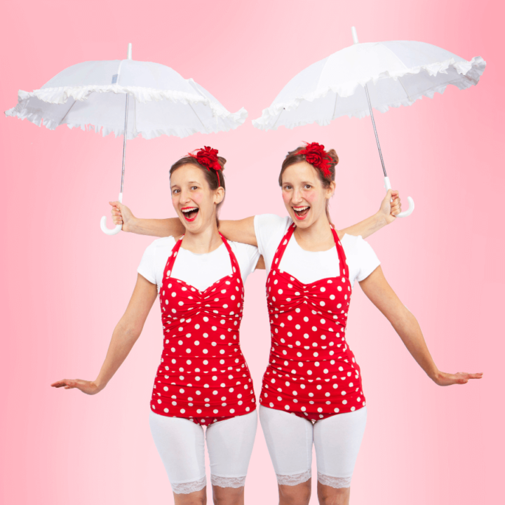 Twin women in red polka dot bathing suits holding white umbrellas.