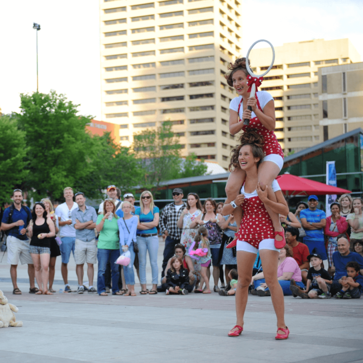 Twin female performers in red polka dot bathing suits perform outside in front of a crowd. One is sitting on the other's shoulders holding a racket.