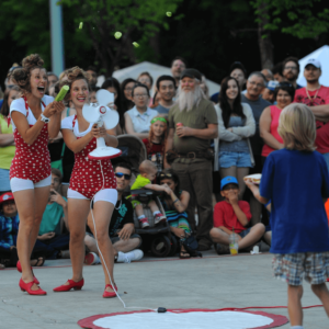 Twin female performers in red polka dot bathing suits perform outside in front of a crowd. They are putting a cucumber through a table fan.