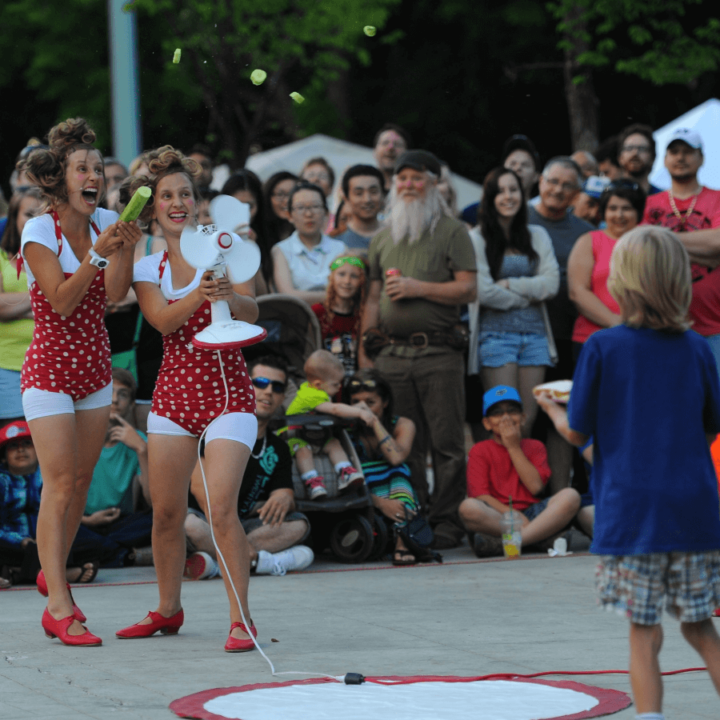 Twin female performers in red polka dot bathing suits perform outside in front of a crowd. They are putting a cucumber through a table fan.