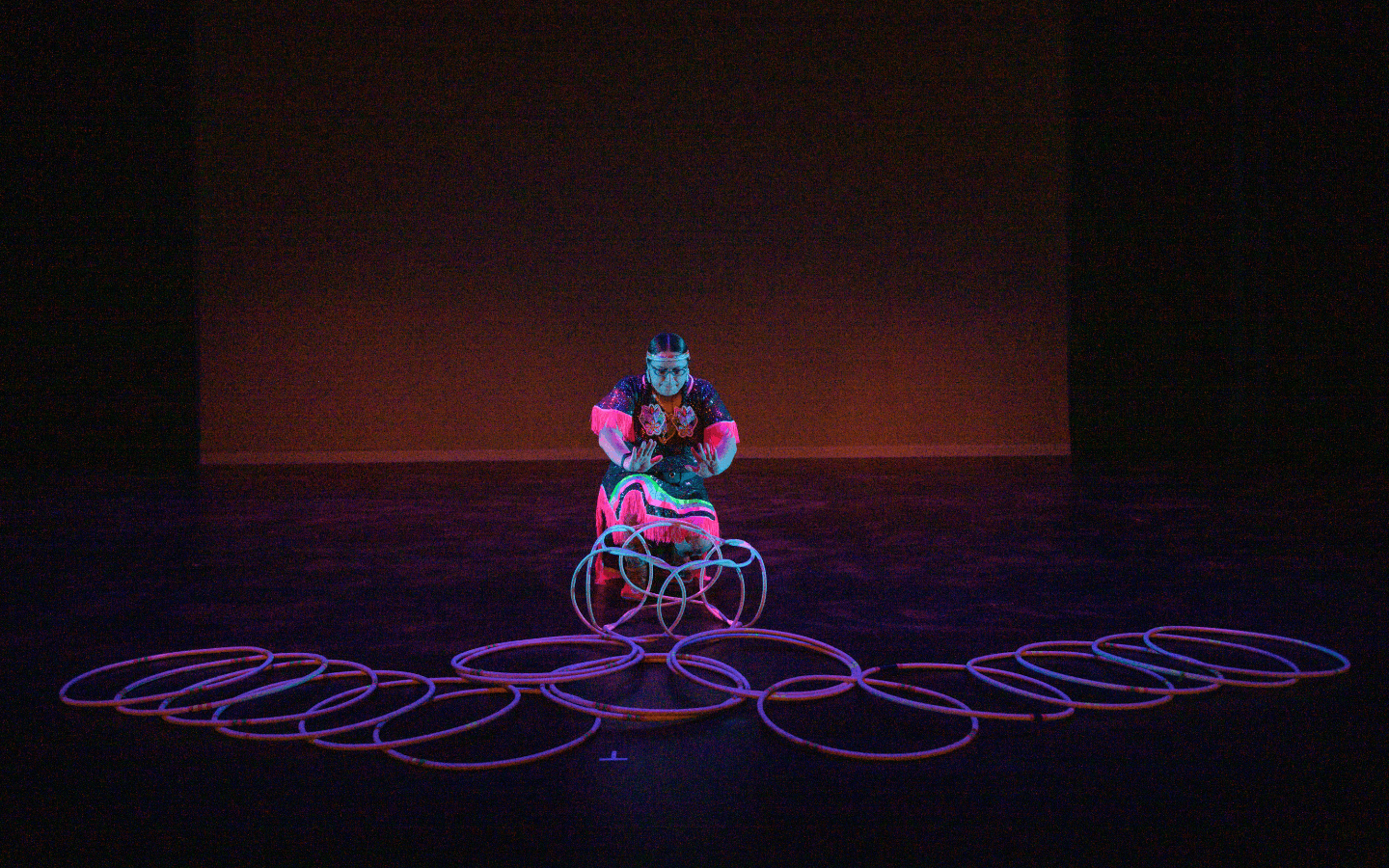 An Indigenous female dancer in traditional regalia crouching on stage with glowing hoops placed in front of her.