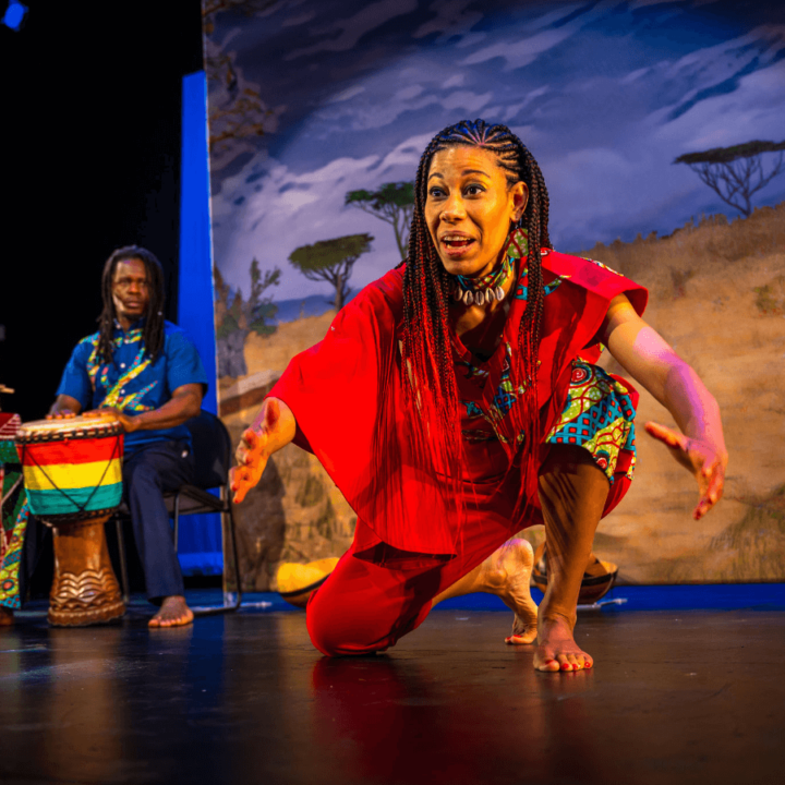 A woman crouches down with her arms out to the audience while telling a story. There's a musician behind her playing a drum. Projected on a screen behind them is an African savannah scene.