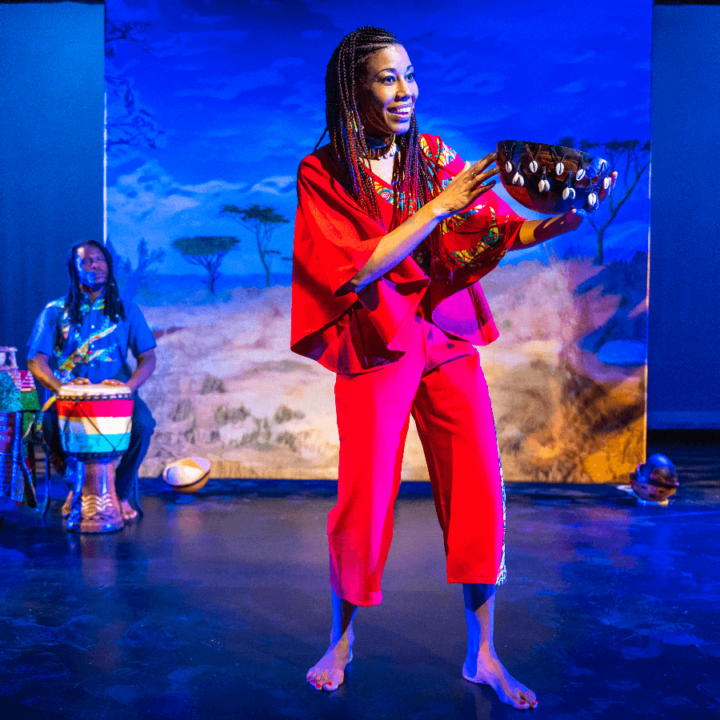 A woman is holding a calabash. There's a musician behind her playing a drum. Projected on a screen behind them is an African savannah scene.