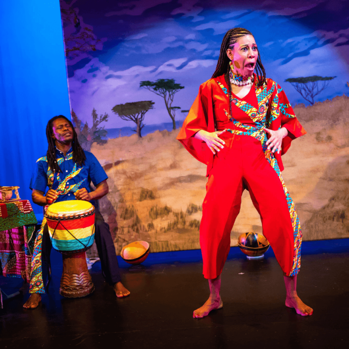 A woman is making a funny pose while telling a story. There's a musician behind her playing a drum. Projected on a screen behind them is an African savannah scene.
