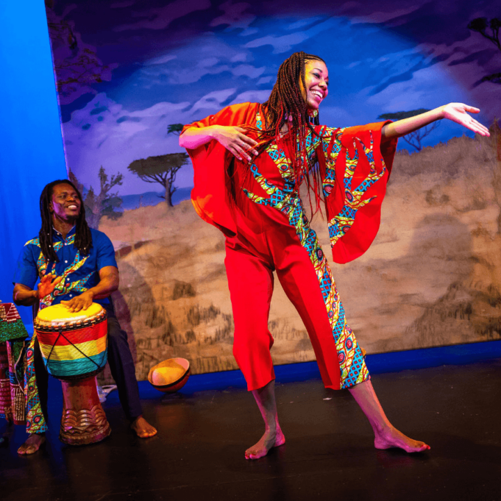 A woman is dancing. There's a musician behind her playing a drum. Projected on a screen behind them is an African savannah scene.