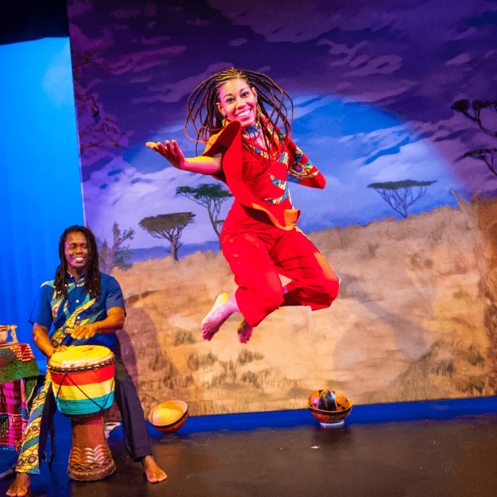 A woman is jumping in the air on a stage. There's a musician behind her playing a drum. Projected on a screen behind them is an African savannah scene.