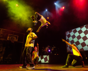A man doing a back flip on stage. A another man is crouching beside him pointing.