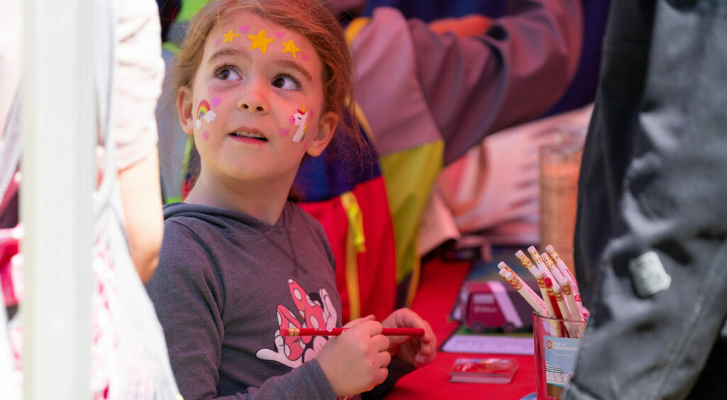 A child looks stands by an activity table, holding a pencil and looking to the left. Her forehead is painted with pink hearts and yellow stars; her left cheek has a rainbow and pink hearts; her right cheek has a unicorn and pink hearts.