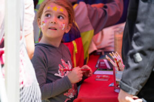 A child looks stands by an activity table, holding a pencil and looking to the left. Her forehead is painted with pink hearts and yellow stars; her left cheek has a rainbow and pink hearts; her right cheek has a unicorn and pink hearts.