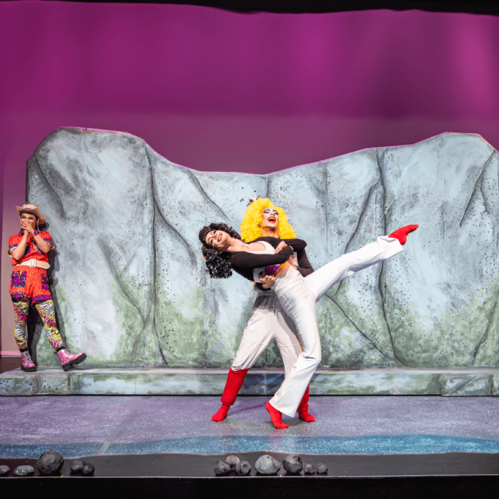 Two performers dancing with each other. One performer has bright yellow, curly hair and the other performer has black hair. They are both wearing black and white jumpsuits and red boots. A third performer, wearing a brightly-coloured safari outfit and a tan hat, is in the background giggling.