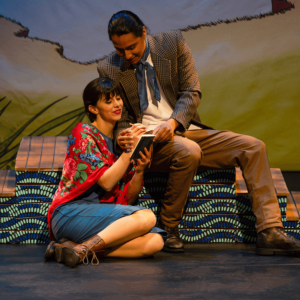 Rose sits on the floor beside. They’re holding a notebook together. He is holding her hand as if showing her how to write.