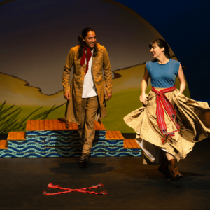 Louis has a red handkerchief tied around his neck. He and Marie are dancing the Red River Jig. Marie’s skirt is flowing. Two red sashes make an X on the floor in front of them.