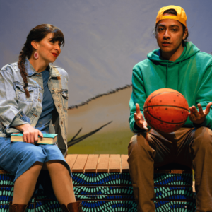 Rose Marie wears a jean jacket over a blue dress. She is sitting, holding a book, and looking at a Métis boy in a hoodie playing with a basketball. It looks like she really wants to kiss him.