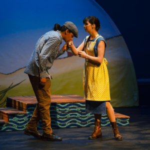 Marie Rose wears a yellow apron over a blue sleeveless dress. Jean, a Métis man wearing a flat cap, is kissing Marie Rose’s hand but she looks worried. This actor plays all the boys and men in the show.