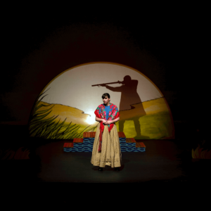 Marie, a Métis woman, is wearing a long mustard yellow skirt and has a floral kokum scarf wrapped around her shoulders. All the girls and women in this show are called Marie or Rose. They are all Métis and played by the same actor. Behind her is a meadow and the shadow of a man pointing a rifle.