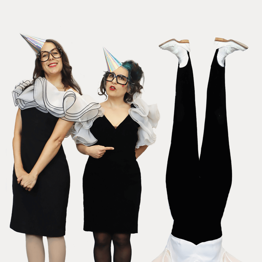 Two white women wearing thick framed glasses, holographic cone-shaped hats, and white ruffles around their shoulders. A pair of legs with white loafers and black tights sticks up in the air next to them.