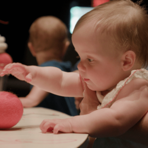 A white baby reaches for a red felt ball.