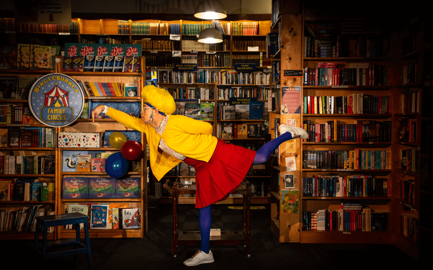 The “libravian,” Lynn, is balancing on one foot and leaning forward like a flamingo. She is wearing a bright yellow cardigan, a red skirt, purple tights, and white sneakers. Her poofy yellow wig matches her cardigan. She is in front of many shelves of books.