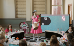 Dottie, the server, is a white woman with short hair wearing a bright pink 1950s-style waitress uniform. She’s speaking with babies and their parents seated around a low circular counter.